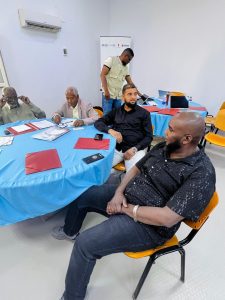 69B5CE6D-7F71-4CBE-9994-E28748FE3732 - جامعة سبها | Sebha University Group of men seated around a round table with blue tablecloth, documents, and folders during a meeting or workshop in a bright room, one person standing in the background.