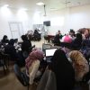 Group of people in a classroom, many women wearing hijabs, working on laptops around tables during a workshop.