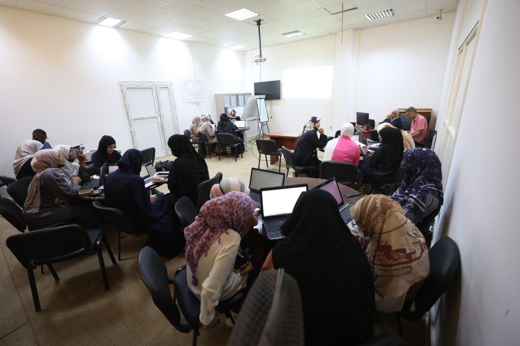 6B54F783-90F8-4F97-9993-38CDC1E8D885 - جامعة سبها | Sebha University Group of people in a classroom, many women wearing hijabs, working on laptops around tables during a workshop.