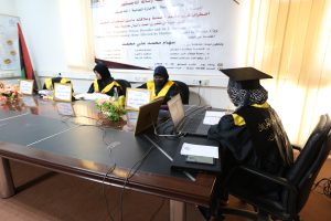 Women in black gowns with yellow sashes sit at a long conference table with laptops, flowers, and framed name cards; a large Arabic banner hangs behind them in a bright room.