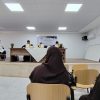 Audience in brown hijabs seated in a classroom watching a panel at the front of a room with a long table and flower arrangements.