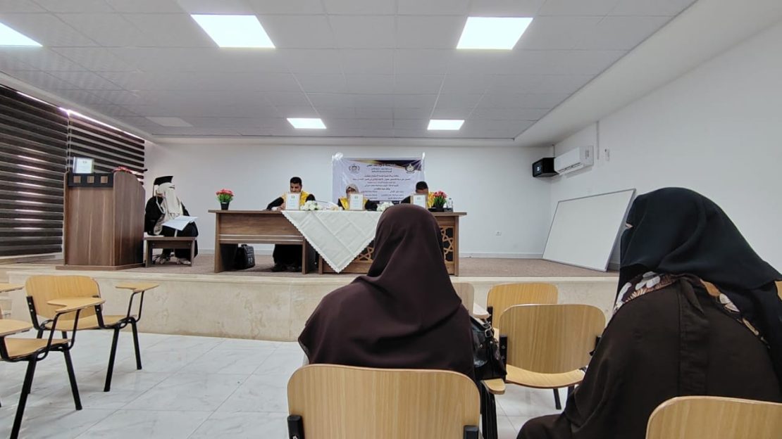 Audience in brown hijabs seated in a classroom watching a panel at the front of a room with a long table and flower arrangements.