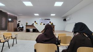 Audience in brown hijabs seated in a classroom watching a panel at the front of a room with a long table and flower arrangements.