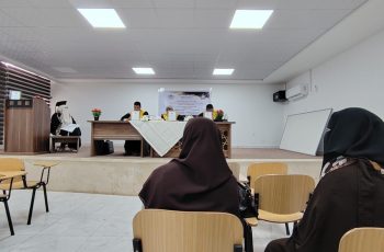 Audience in brown hijabs seated in a classroom watching a panel at the front of a room with a long table and flower arrangements.