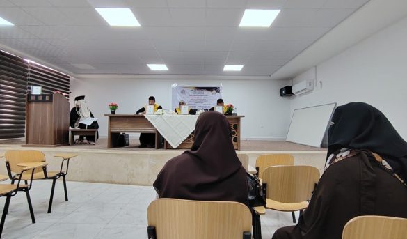 Audience in brown hijabs seated in a classroom watching a panel at the front of a room with a long table and flower arrangements.