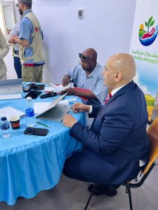 Two men sit at a blue table with a laptop, papers, and water bottles, while a banner for the Sawaed Project is in the background.