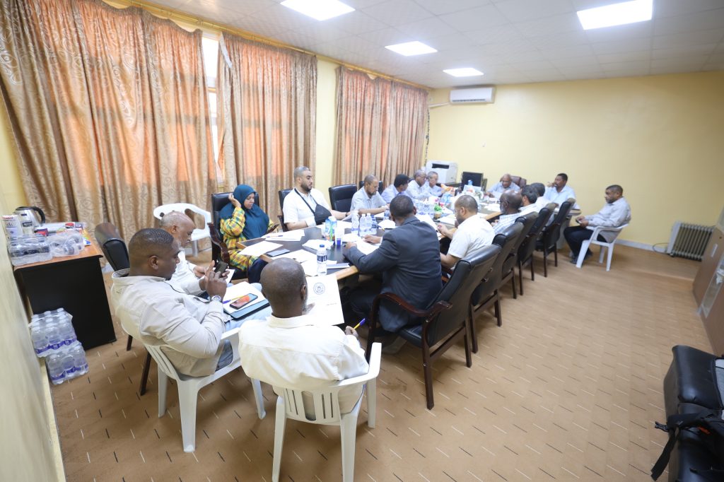 704426F0-19FC-4CB1-A0BB-710BEA72ADB8 - جامعة سبها | Sebha University A long conference table with about twenty people seated around it, discussing papers and using laptops in a bright room with beige walls and curtains.