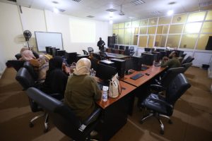 71F343D1-C301-4877-B413-514A09883825 - جامعة سبها | Sebha University Instructor presents to students in a computer lab, standing at the front as a projection screen and whiteboard are visible behind them.