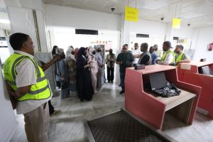 Group of visitors listening to a guide at a security checkpoint with red kiosks and staff in hi-visibility vests.