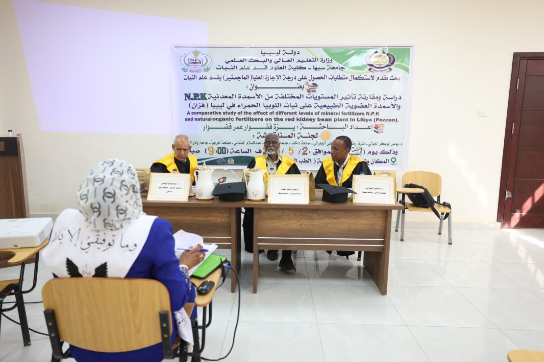 Three men in academic gowns seated at a table on a podium, facing an audience in a classroom, with a large banner in Arabic and English behind them.