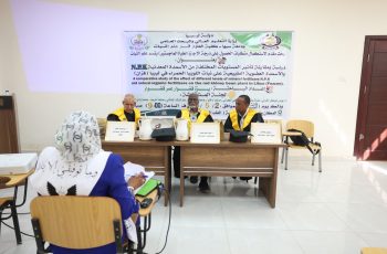 Three men in academic gowns seated at a table on a podium, facing an audience in a classroom, with a large banner in Arabic and English behind them.