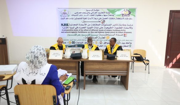 Three men in academic gowns seated at a table on a podium, facing an audience in a classroom, with a large banner in Arabic and English behind them.