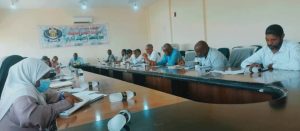 People sit around a large U-shaped conference table in a meeting room, banner with Arabic text on the wall behind them.