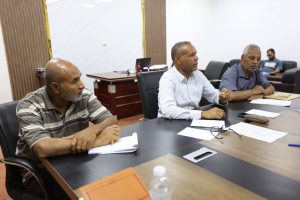 790F3F7A-0D94-4A2E-81FE-0ED2D235D300 - جامعة سبها | Sebha University Four men in a meeting room sit around a conference table with papers, discussing a project.