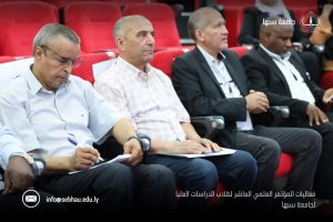 794BB128-EB9E-4FDF-9112-2889EF9B269F - جامعة سبها | Sebha University Men in business-casual attire sit in red auditorium seats at a conference, some taking notes, with Sebha University branding in the corner.