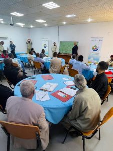 A training workshop in a conference room with attendees seated around round tables covered in blue cloths, facing a presenter at the front with a large screen and banners in the background.