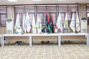 Two officials sit at a long conference table in a formal meeting room, flags displayed behind them, including a Libyan flag center-back.