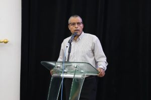 85A42820-69E4-426D-8B88-771014D5FCCC - جامعة سبها | Sebha University Man in a striped shirt speaks at a clear glass podium on a stage with a black curtain backdrop.