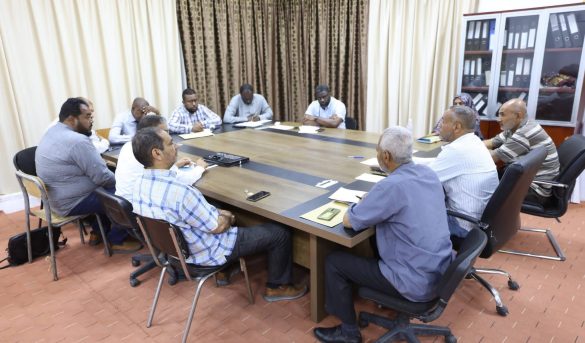 85DC208A-B319-44DF-BE41-AABD272D3AE5 - جامعة سبها | Sebha University A diverse group of people seated around a large conference table, talking and taking notes in a meeting room with files on a cabinet in the background.