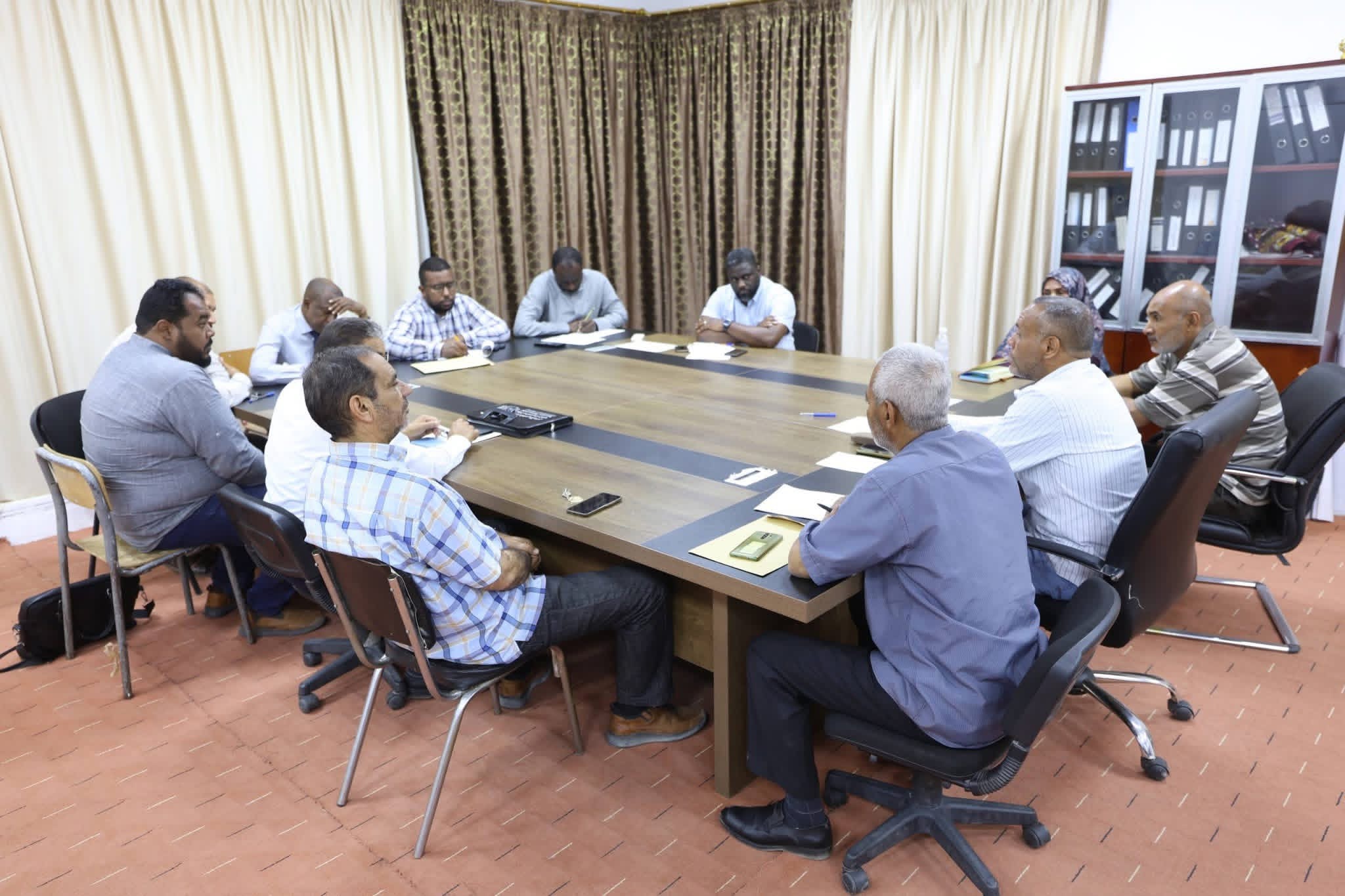 A diverse group of people seated around a large conference table, talking and taking notes in a meeting room with files on a cabinet in the background.
