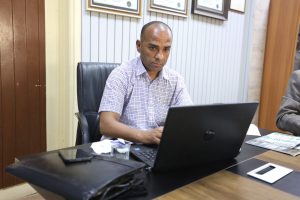 Man in a checkered shirt seated at a desk, looking at a laptop in a tidy office with framed certificates on the wall behind him.