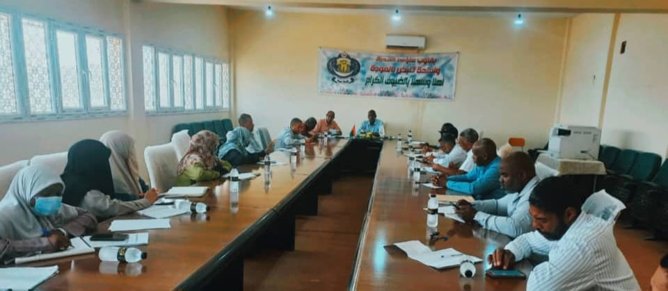 Diverse group of people seated around a long conference table, taking notes during a meeting in a bright room with a banner on the back wall.