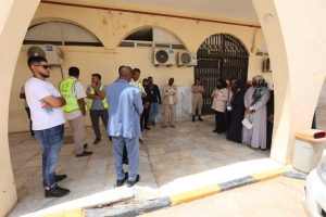 Group of people with reflective vests and others in casual attire stand in a sunlit courtyard under arches outside a beige building with air conditioners.",