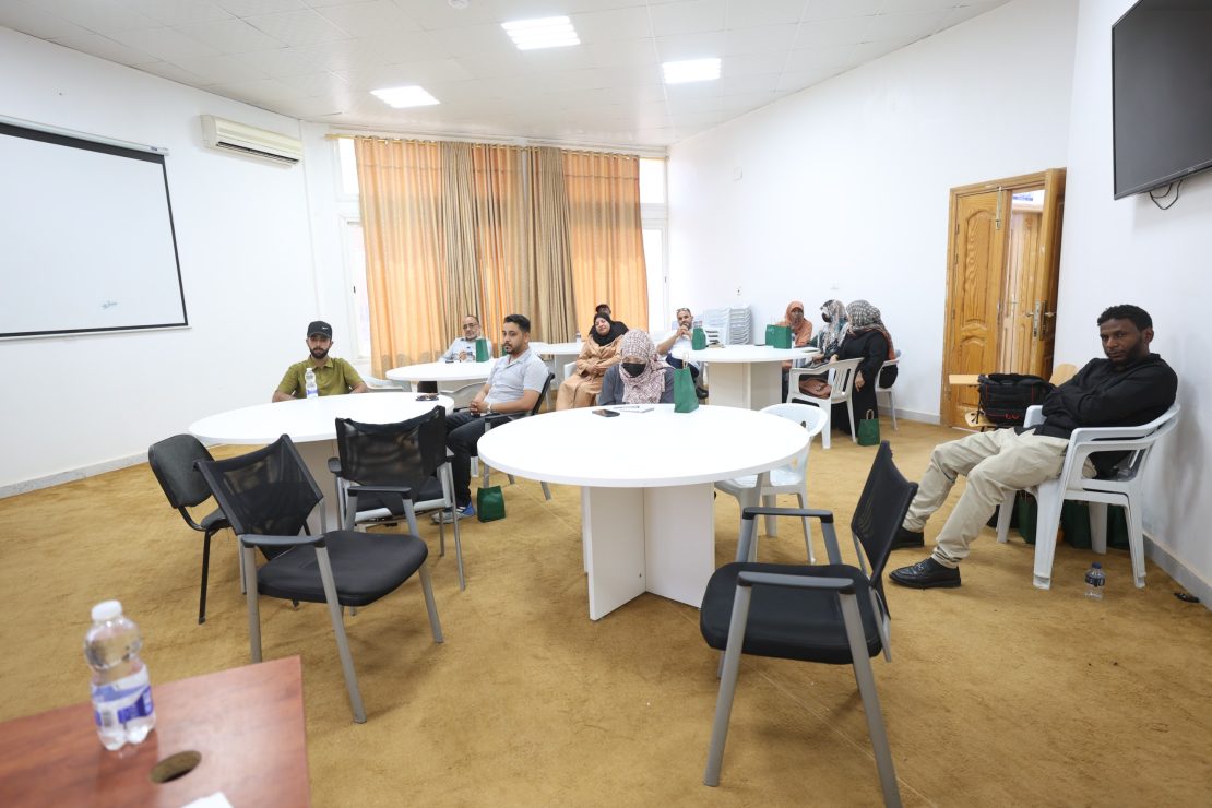 A conference room with round tables and people seated, some with notebooks and bags, beige carpet, cream curtains, and a wall-mounted TV on the right.