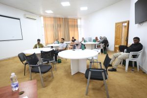 9371722E-1721-4525-95AD-D20D03D04E4F - جامعة سبها | Sebha University A conference room with round tables and people seated, some with notebooks and bags, beige carpet, cream curtains, and a wall-mounted TV on the right.