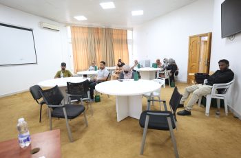 A conference room with round tables and people seated, some with notebooks and bags, beige carpet, cream curtains, and a wall-mounted TV on the right.