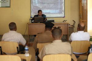 Speaker at a desk giving a lecture to students in a classroom, with laptop and projector behind him