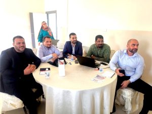 Group of six adults seated around a round table in a bright room, one woman in a hijab standing near a doorway in the background.