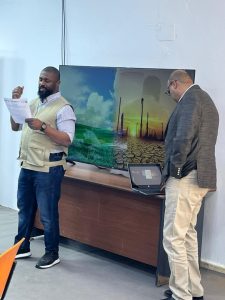 Two men presenting beside a TV showing a split image of green fields and a cracked desert with oil rigs, one holding papers while the other looks at a laptop.