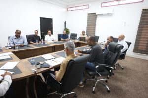 9C8A2169-F0A2-4632-9B89-69D0D2320211 - جامعة سبها | Sebha University Group of professionals seated around a large wooden conference table in a bright meeting room, with papers and water bottles on the table.
