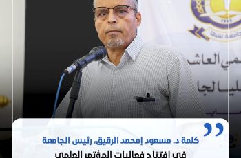 A middle-aged man wearing glasses speaks at a podium with a microphone, backdrop shows a banner and crest.