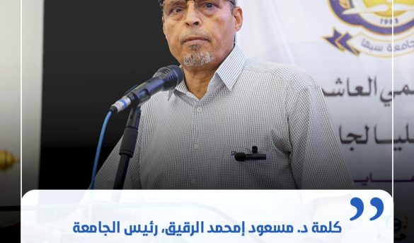 9DF81B0F-A665-4173-8177-0F9DE6DD10BA - جامعة سبها | Sebha University A middle-aged man wearing glasses speaks at a podium with a microphone, backdrop shows a banner and crest.