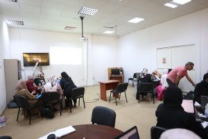 A7136611-29CC-49C9-A934-89225C2DF09A - جامعة سبها | Sebha University Group of women in hijabs seated around tables in a classroom, with a projector screen visible at the front and a few laptops open on desks to the right.