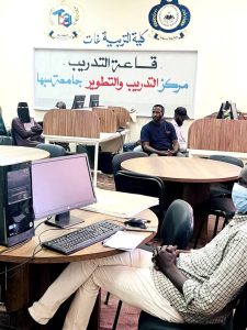 A computer lab at a women’s college with several desktop PCs, desks, and students seated, some wearing traditional head coverings, sign in Arabic on the wall behind.