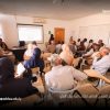 Large classroom seminar with a speaker at the front presenting a projected slide to a full audience of adults; orange curtains on the left and a projector screen visible. “جامعة سبها” logo appears on the top right.