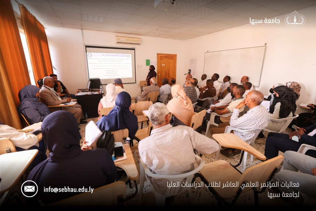 Large classroom seminar with a speaker at the front presenting a projected slide to a full audience of adults; orange curtains on the left and a projector screen visible. “جامعة سبها” logo appears on the top right.