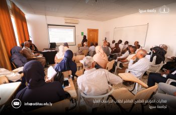 Large classroom seminar with a speaker at the front presenting a projected slide to a full audience of adults; orange curtains on the left and a projector screen visible. “جامعة سبها” logo appears on the top right.