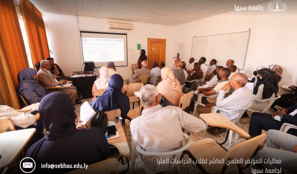 AC84E642-9D69-496E-B134-F9B333E9E823 - جامعة سبها | Sebha University Large classroom seminar with a speaker at the front presenting a projected slide to a full audience of adults; orange curtains on the left and a projector screen visible. “جامعة سبها” logo appears on the top right.