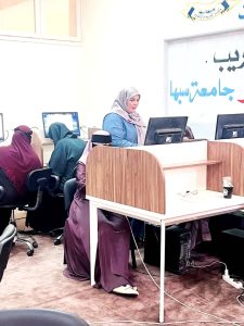 A woman in a blue top and light hijab stands at a computer station in a classroom, with students at computers in the background.