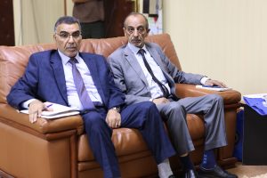B28C5A41-CD79-49FD-87EB-C7872FDBBC38 - جامعة سبها | Sebha University Two men in suits sitting on a brown leather sofa, looking at the camera in a office lounge setting.
