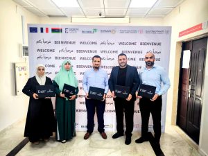 Five people pose together indoors in front of a 'Bienvenue Welcome' backdrop, each holding dark folders.