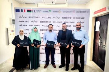 Five people pose together indoors in front of a 'Bienvenue Welcome' backdrop, each holding dark folders.