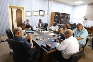 Group of men seated around a long conference table in a meeting room, some with laptops and papers, in discussion.