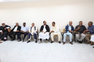 Group of men in traditional and formal attire sitting on chairs with attached desks along a wall, likely in a meeting or discussion.