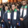 Group of men in suits standing at a formal ceremony, wearing white scarves with green and red Algerian flags; audience seated behind.