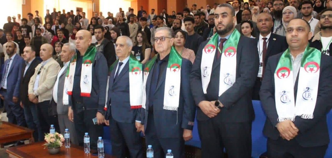 Group of men in suits standing at a formal ceremony, wearing white scarves with green and red Algerian flags; audience seated behind.